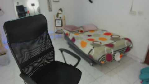 Media: A video of a small, sparsely furnished bedroom with a black office chair in the foreground, a single bed with a colorful, patterned bedspread, and minimalistic decor.