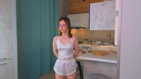 Video of a slender young woman with straight, shoulder-length brown hair, wearing pink glasses, a white camisole, and matching shorts, standing in a modern kitchen with light wood cabinets and a teal curtain.
