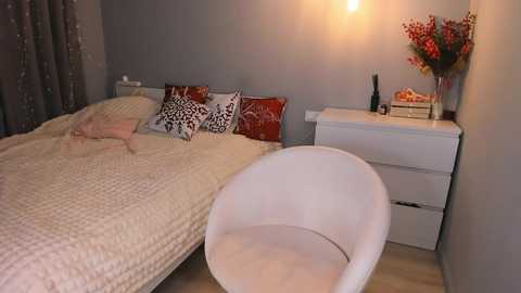 Video of a modern bedroom with a beige quilted bed, red and white patterned pillows, a white dresser, a white chair, and a vase of red flowers.
