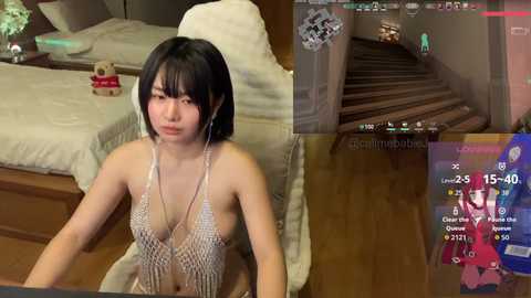 Media: A video featuring an Asian woman with short black hair, wearing a revealing silver halterneck dress, sitting on a bed in a modern bedroom with wooden floors and a gaming screen displaying a virtual staircase.