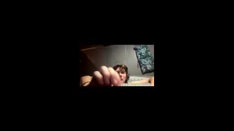 Video of a boy with curly brown hair, lying on a bed, looking towards the camera. The room has a wooden headboard, a floral-patterned wall hanging, and dim lighting.