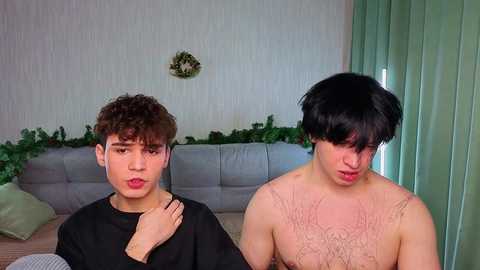Video of two young men: one with curly brown hair, wearing a black sweater, the other with short black hair, shirtless, in a room with green curtains and a gray couch.