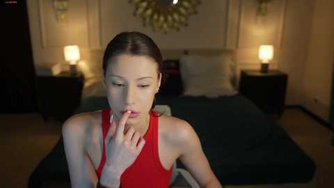Media: Video of a young Asian woman with fair skin, dark hair, and a slender physique, wearing a red sleeveless top, sitting pensively on a bed in a dimly lit bedroom with two bedside lamps and a floral wall hanging.