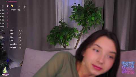 A video of a young woman with straight brown hair and light skin, wearing a green top, leaning forward, in a dimly lit living room with green curtains and a potted plant.