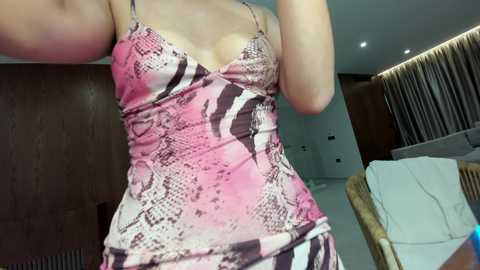 A video of a woman in a tight, pink and black snake-print dress, indoors, with wooden paneling, a white couch, and a wooden chair in the background.