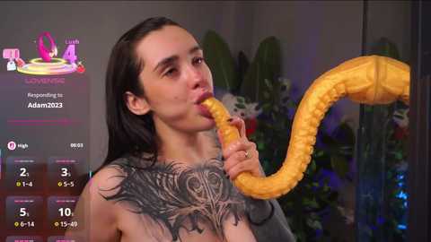 Video of a young woman with long black hair, wearing a gray top, eating a large, yellow, cartoon-like octopus with tentacles. Background features green plants.
