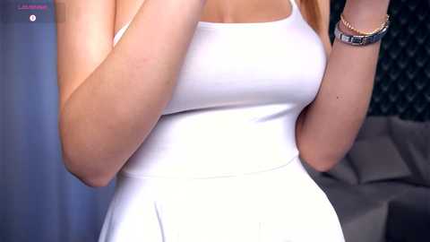 Video of a fair-skinned woman in a white, sleeveless dress, holding her arms up. She has a silver wristwatch and wears a bracelet. Background features a dark patterned wall and a gray couch.