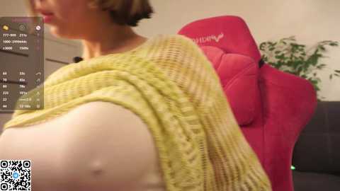 A video captures a partially visible, fair-skinned woman with a short brown bob, wearing a yellow knit sweater. She sits in a red gaming chair with a potted plant in the background.