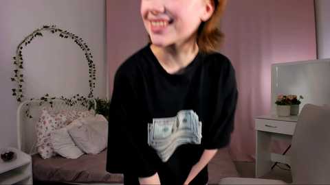 Video of a smiling young woman with fair skin and reddish-brown hair, wearing a black T-shirt with a dollar bill graphic. Background includes a white bed with pillows, a white desk, and a pink wall.