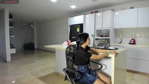 Video of a modern kitchen with a woman in a black top and denim skirt seated at a counter, facing away, typing on a laptop. White cabinets, stainless steel appliances, and light beige tiled floor.
