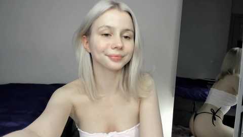 Video of a young, fair-skinned woman with platinum blonde hair, wearing a strapless white top, standing in a dimly lit room with a dark blue bed in the background.