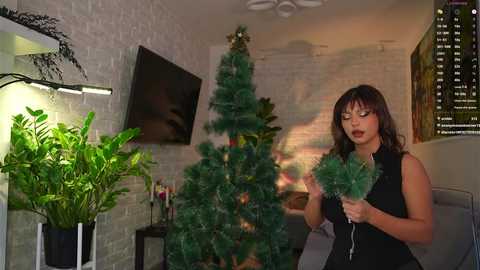 Video of a woman with medium skin tone and long brown hair, wearing a black dress, decorating a fake Christmas tree with green ornaments in a modern, dimly lit living room.