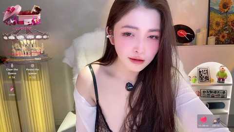 Video of a young Asian woman with long brown hair, fair skin, wearing a black lace bra and white robe, seated in a cozy room with colorful decor.