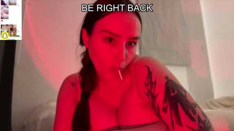 Video of a tattooed woman with a cigarette in her mouth, wearing a strapless top, under red lighting, with the text \"BE RIGHT BACK\" above her.