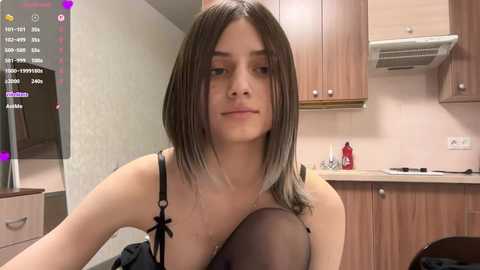 Video of a young woman with straight, shoulder-length brown hair in a kitchen, wearing a black bra and a black top, with wooden cabinets and a stove in the background.