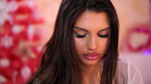 Video of a young woman with long, dark hair, wearing dark eye makeup and a white shirt, standing in front of a blurred red and white background.