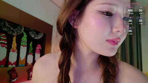 Video of a young Caucasian woman with light skin and long brown hair in braids, wearing a red top. Background includes a Christmas-themed wall with festive decorations.