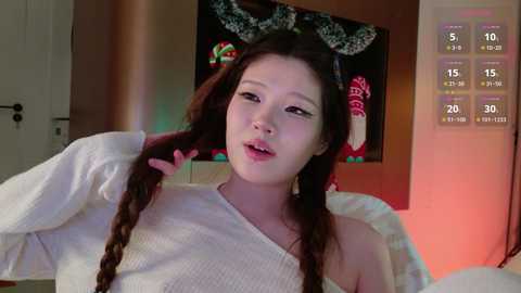 Video of a young Asian woman with long, dark braided hair, wearing a white off-shoulder knit sweater, sitting on a bed. Background features holiday decorations and a video game screen displaying stats.