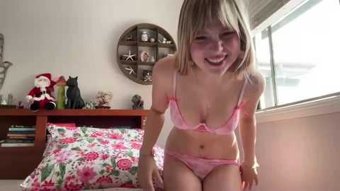 A video of a smiling young woman with fair skin and straight blonde hair, wearing pink lace lingerie, standing in a sunlit bedroom with a decorative shelf, a Christmas elf, and floral bedding.