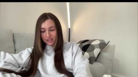 Video of a young Caucasian woman with long, straight brown hair, wearing a light gray sweatshirt, sitting on a light gray couch in a modern living room.