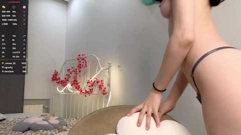 Video of a nude woman with light skin, wearing a green hat, standing near a bed with a white metal headboard adorned with red flowers. The room is dimly lit, and a live streaming interface with stats is visible in the upper left corner.