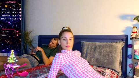 Video of a young woman with light skin and brown hair, wearing a pink houndstooth sweater, lying on a gray bed with a man behind her, both in a cozy, dimly lit bedroom with a nightstand and cityscape view.