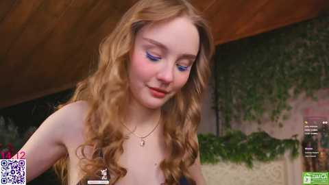 A video of a young, fair-skinned woman with wavy, light brown hair, wearing blue eyeshadow, a black strapless dress, and a gold necklace, standing in a rustic, wooden-beamed room with a green ivy wall.