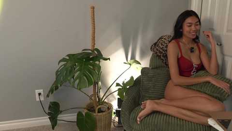 A video shows a slender, light-skinned Asian woman with long black hair, wearing a red bra and panties, sitting nude on a green chair. She's smiling, with a potted plant beside her.