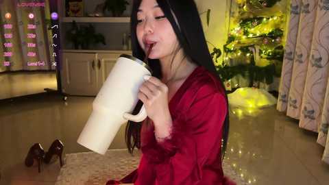 A video of an Asian woman with long black hair, wearing a red satin robe, sitting on the floor, drinking from a white mug, surrounded by a festive, warmly lit living room with a decorated Christmas tree.