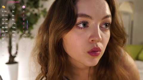 Video of a young woman with wavy brown hair, light skin, and pink lipstick, looking pensive. Background includes a blurred green plant and white wall.
