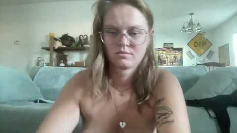 Media: Video of a fair-skinned, middle-aged woman with glasses, topless, sitting on a couch, wearing a necklace with a heart pendant, in a living room with a blue couch, shelves, and a yellow \"DIP\" sign on the wall.