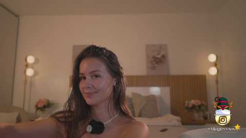 Video of a woman with wet hair, wearing a black necklace, standing in a warmly lit bedroom with wooden headboard, floral wallpaper, and twin bedside lamps.