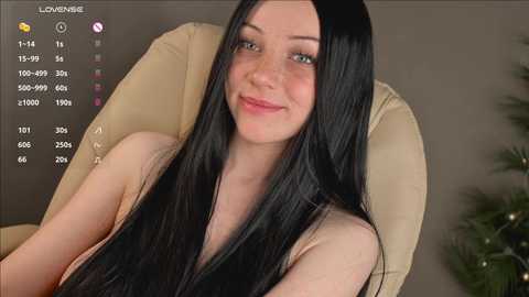 Video of a fair-skinned woman with long black hair, freckles, and blue eyes, smiling. She is topless, sitting in a beige chair against a plain brown wall.