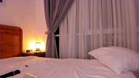 Video of a cozy bedroom with a wooden headboard, white bed, and sheer curtains. Nightstand with a lamp and remote control.