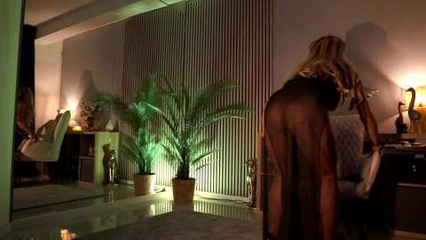 Video of a blonde woman in a sheer black bodysuit, bending over in a dimly lit, modern room with green plants, a mirror, and a white chair.