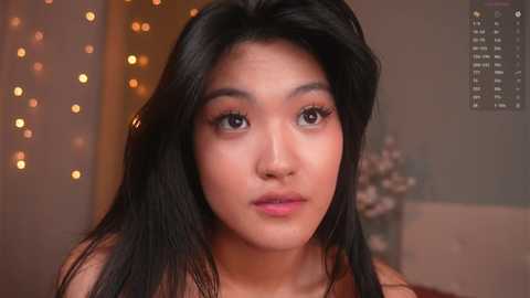 Video of an Asian woman with long, straight black hair and light skin, wearing makeup, looking serious, in a dimly lit room with warm lighting and blurred bokeh effects.