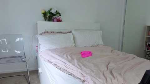 A minimalist, modern bedroom photo featuring a neatly made bed with white sheets, a pink blanket, and a vibrant pink pillow. A clear acrylic chair is on the left, and a white bookshelf on the right.