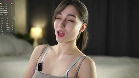 Video of a young woman with fair skin, brown hair, wearing a light grey tank top, lying on a bed with a blurred, dark background. She has a relaxed expression with her mouth slightly open.
