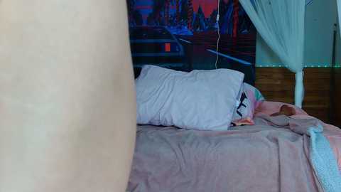 Video of a messy bedroom with a beige bed, white pillows, and a colorful blanket. A person's leg is partially visible on the left. The background features a large, colorful mural and string lights.