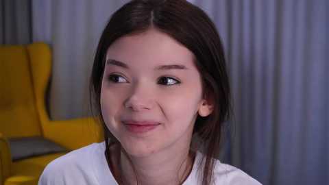Video of a young woman with light skin, dark brown hair, and minimal makeup, smiling softly. She's wearing a white top. Background features a mustard-yellow chair and gray curtains.