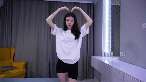 Video of an East Asian woman with straight black hair, fair skin, and a slender physique, making a heart shape with her hands in a minimalist, modern room with grey curtains, a yellow chair, and a white sink.
