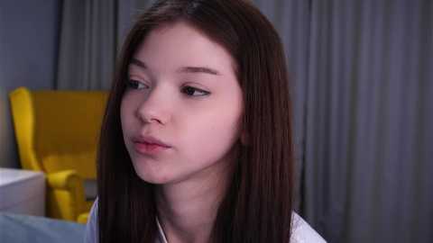 Video of a young Asian woman with straight, shoulder-length dark hair, fair skin, and light makeup. She wears a white shirt, gazing thoughtfully. Background features a yellow chair and gray curtains in a modern, indoor setting.