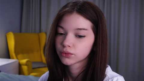Video of a young girl with long, straight brown hair, light skin, and a neutral expression, wearing a white shirt, sitting in a modern room with gray curtains and a yellow armchair.