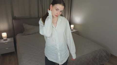 A video of a young Caucasian woman with fair skin and long dark hair, wearing a white button-down shirt and dark pants, standing in a dimly lit bedroom with a bed and two bedside lamps.