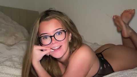 Video of a young Caucasian woman with long blonde hair, wearing glasses and black lingerie, lying on her stomach on a white bed with beige pillows in a simple, dimly lit bedroom.