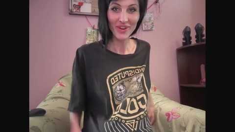 Video of a young woman with pale skin, black hair, and a large, black graphic t-shirt featuring a cartoon character. She sits on a bed with floral bedding, in a pink room adorned with posters and dark figurines.