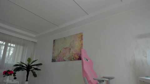 A video of a modern, minimalist room with white walls, a pink recliner chair, and a colorful abstract painting. A potted plant with red flowers is on the left.