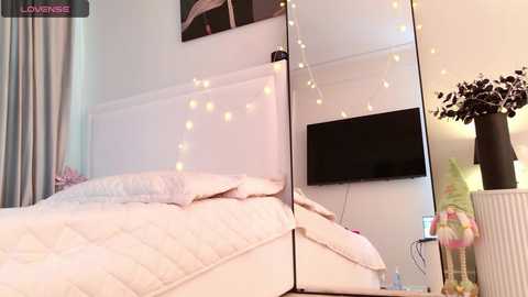 A video of a modern, cozy bedroom with a white, plush bed, a flat-screen TV on the wall, fairy lights, and a vase with faux foliage.