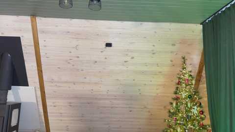 Video of a cozy attic room with light wood paneling, a small Christmas tree adorned with red and gold ornaments, and a green curtain.