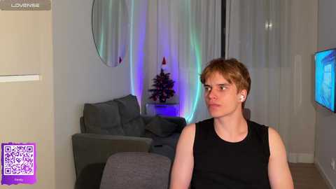 Media: Video of a young white man with short brown hair, wearing a black sleeveless shirt, sitting in a modern living room with a gray couch, TV, and colorful LED lights.